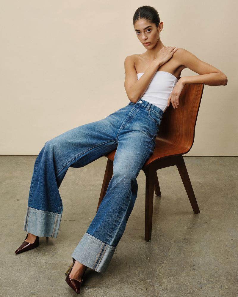 A woman sits on a wooden chair, posing in a white tube top, wide-legged blue jeans, and dark brown heels in a studio shot.