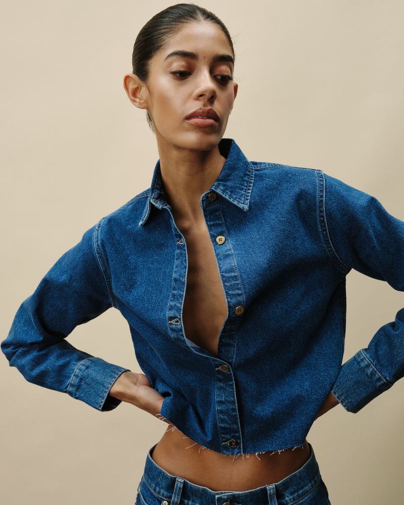 Portrait of a brunette female model in a blue denim collared crop top and jeans against a neutral background in a studio shot.