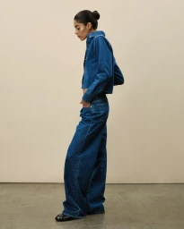Model in dark blue denim shirt and matching wide-leg jeans standing against a warm neutral studio backdrop