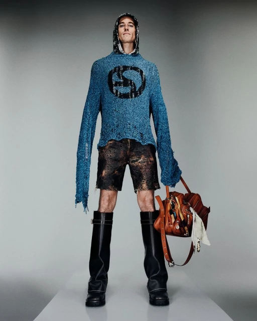 Fashion model in blue hoodie with symbol, brown shorts, and black boots holds a brown leather bag in a full-length studio shot.