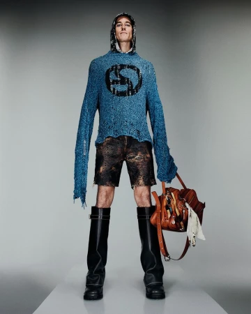 Fashion model in blue hoodie with symbol, brown shorts, and black boots holds a brown leather bag in a full-length studio shot.