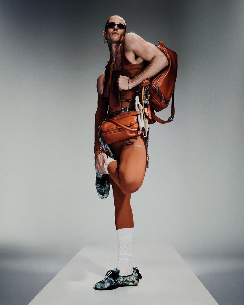 Male fashion model in brown outfit and sunglasses poses with backpack and crossbody bag in a bright studio setting.