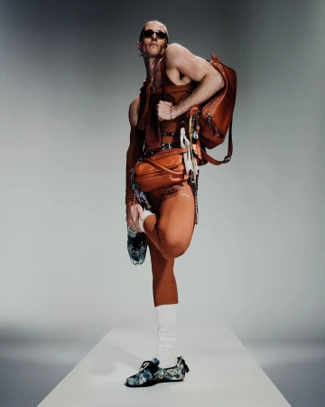 Male fashion model in brown outfit and sunglasses poses with backpack and crossbody bag in a bright studio setting.
