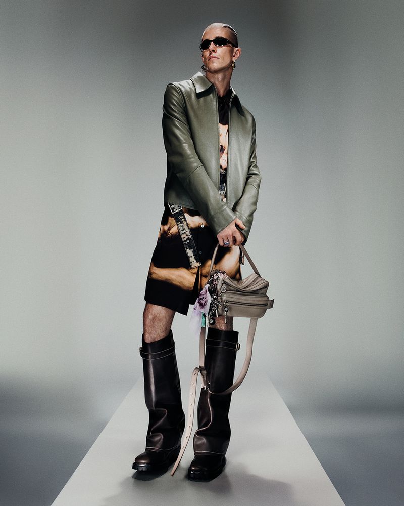 Full-length studio shot of an adult male wearing a jacket, patterned shirt and shorts, sunglasses, and knee-high boots, holding a purse.
