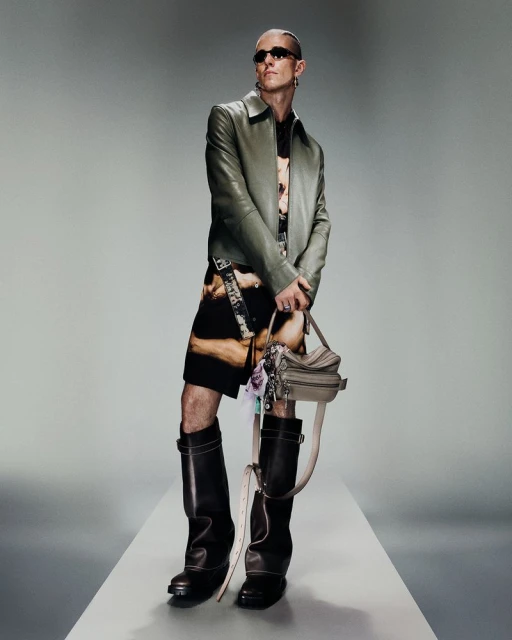 Full-length studio shot of an adult male wearing a jacket, patterned shirt and shorts, sunglasses, and knee-high boots, holding a purse.
