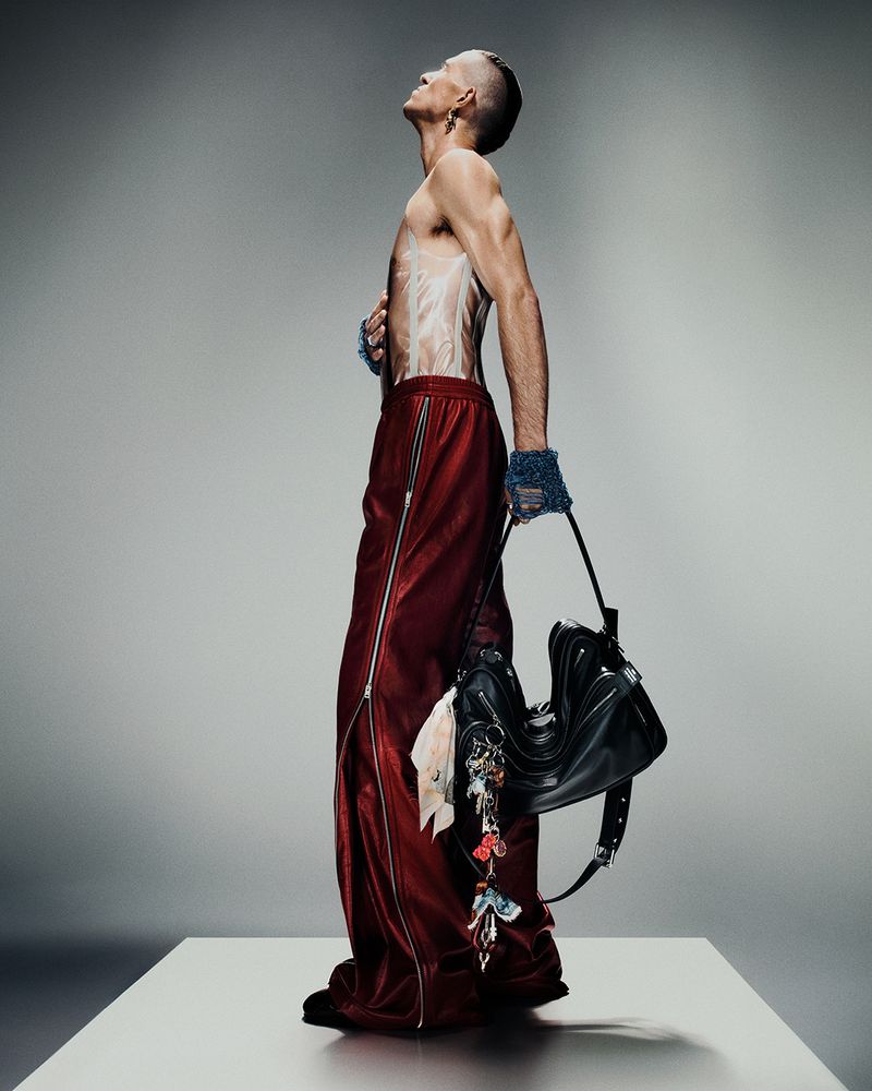 Profile shot of a topless adult male in baggy red pants holding a black bag with small accessories in a gray photo studio.