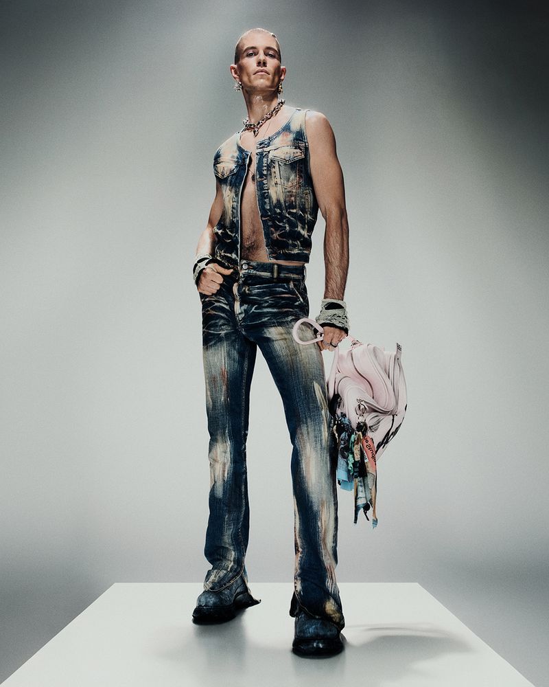 Man with buzzcut poses in distressed denim vest and pants, jewelry and black boots, holding a multi-colored fabric bag in a photo studio.