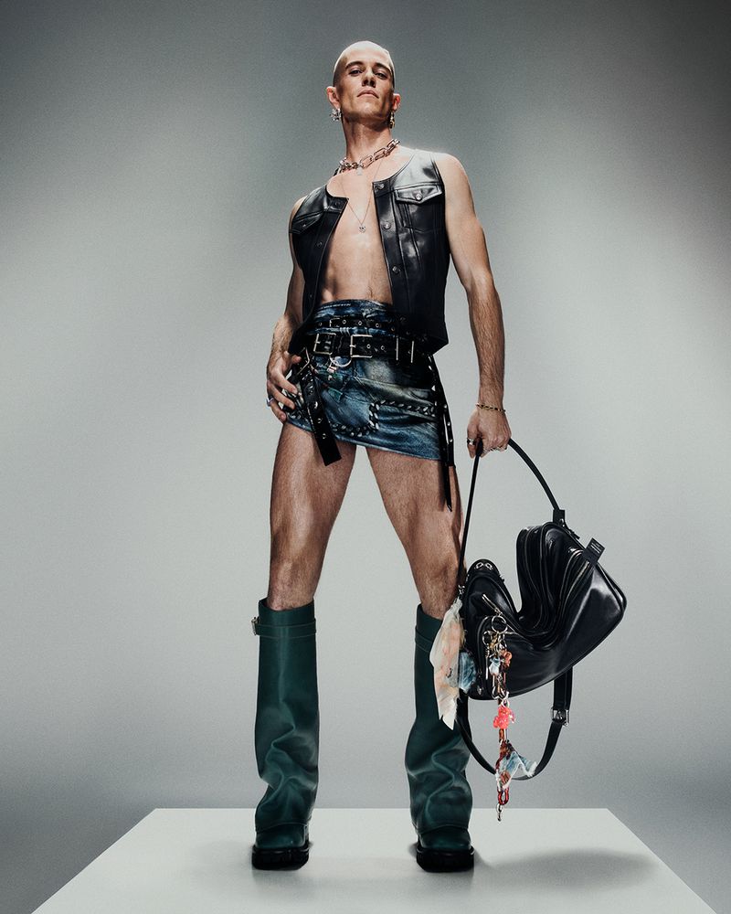 A person stands in a studio wearing a black vest and a denim skirt, holding a black purse with dangling accessories, against a neutral backdrop.