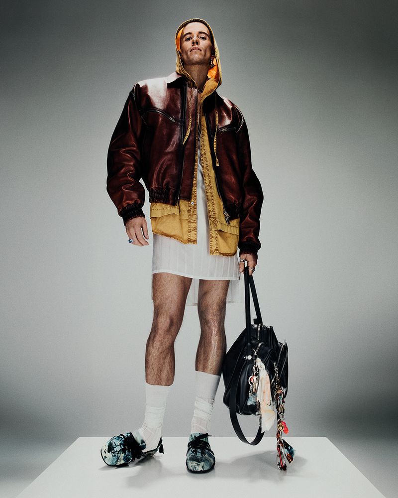 Adult male posing in a studio wearing a brown leather jacket, white dress, socks and heeled shoes, and carrying a black leather bag.