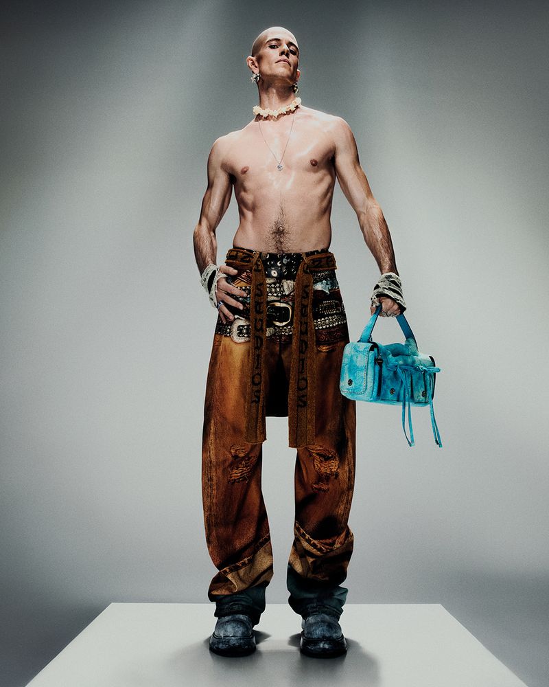 Full-length portrait of a slim, bald adult male with multiple accessories, holding a blue purse, wearing low-slung pants in a studio setting.