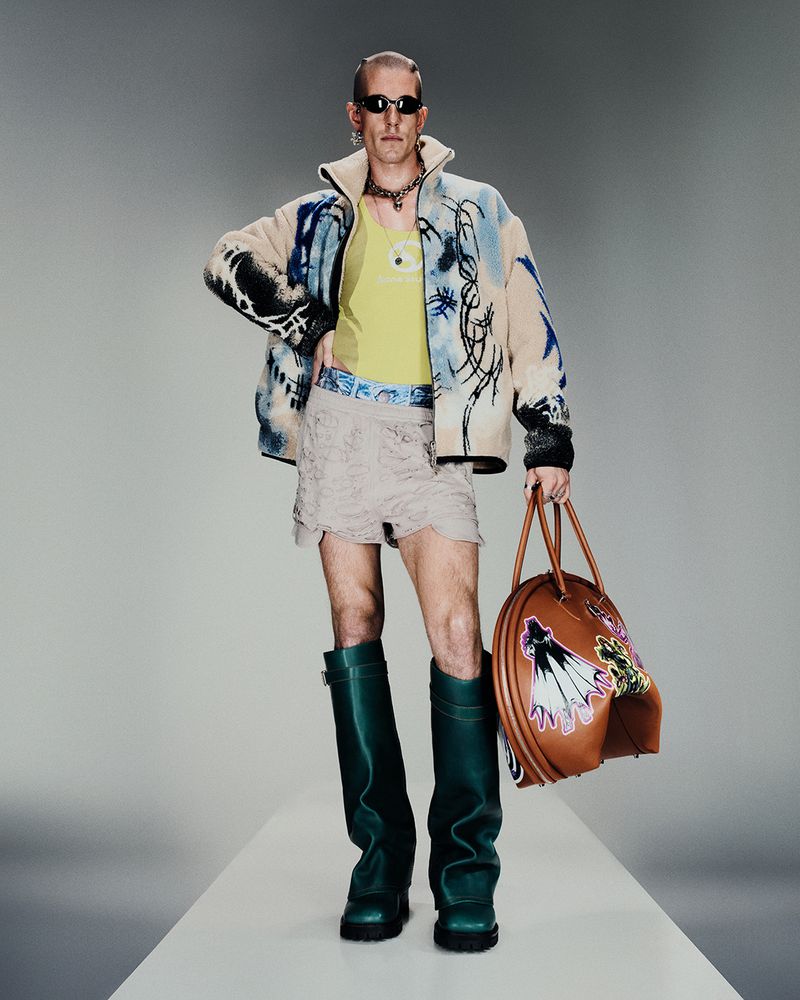 Full-length studio shot of a male fashion model wearing sunglasses, patterned jacket, tank top, lace shorts, boots, and carrying a bag.
