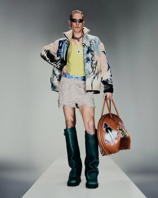 Full-length studio shot of a male fashion model wearing sunglasses, patterned jacket, tank top, lace shorts, boots, and carrying a bag.