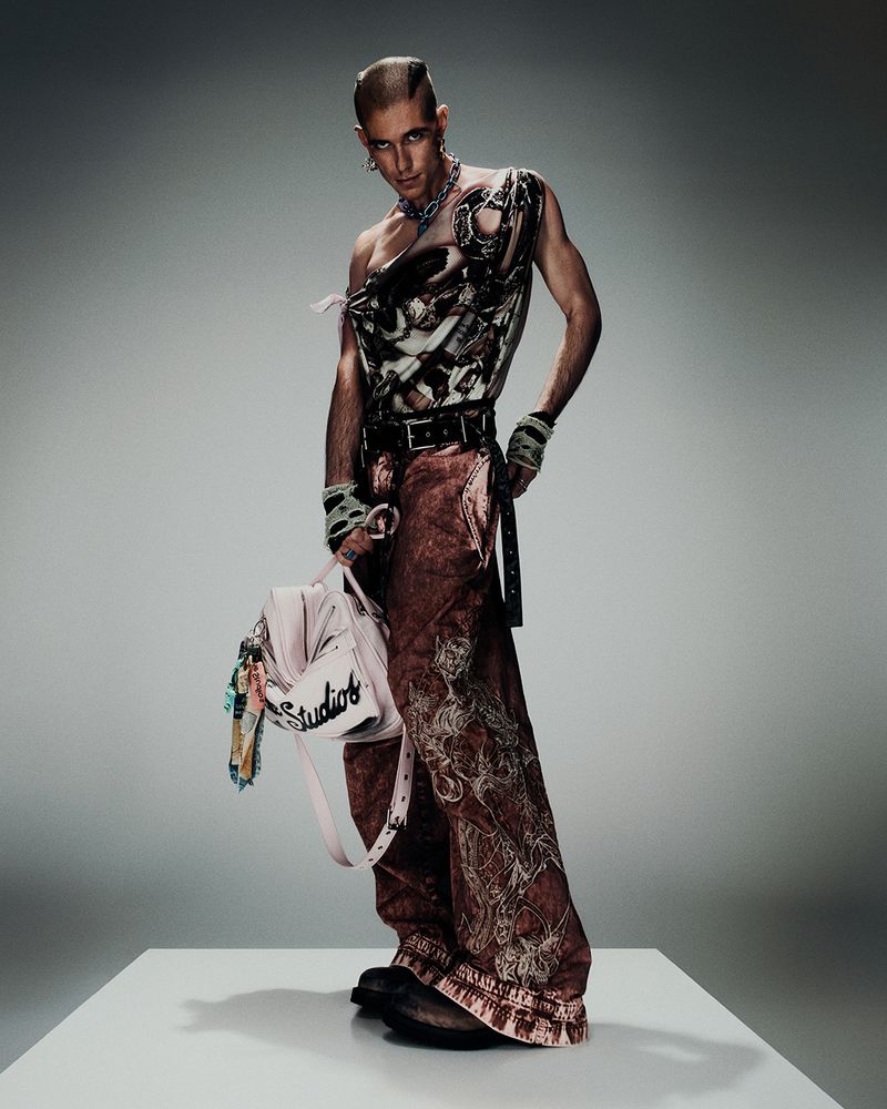 A person with a buzzed head is posing in a sleeveless top and wide-legged pants, holding a purse, standing on a white platform against a gray background.