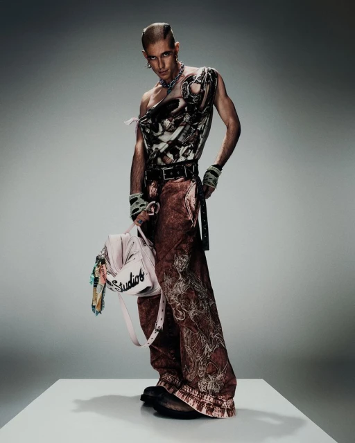 A person with a buzzed head is posing in a sleeveless top and wide-legged pants, holding a purse, standing on a white platform against a gray background.