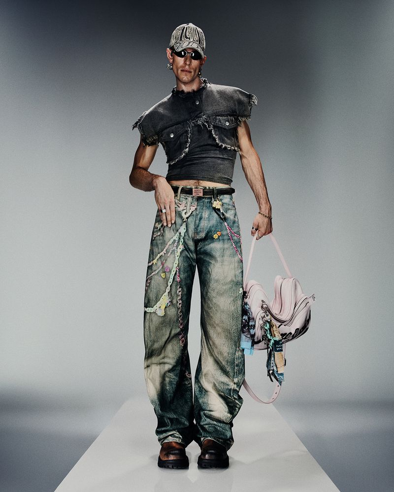 Male model in a dark cropped vest top and patterned jeans holds a pink handbag in a studio shot on a white runway.