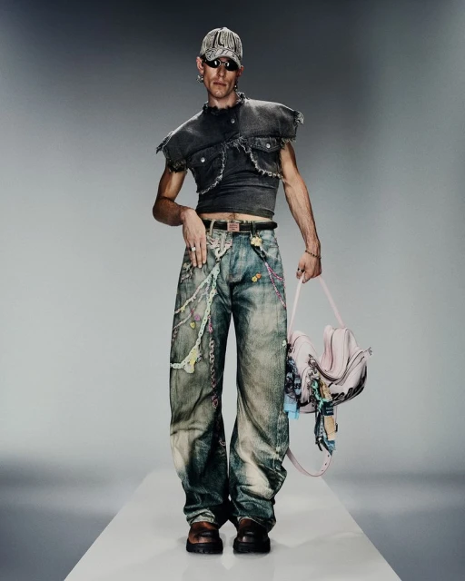 Male model in a dark cropped vest top and patterned jeans holds a pink handbag in a studio shot on a white runway.
