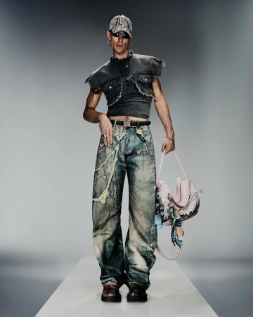 Male model in a dark cropped vest top and patterned jeans holds a pink handbag in a studio shot on a white runway.