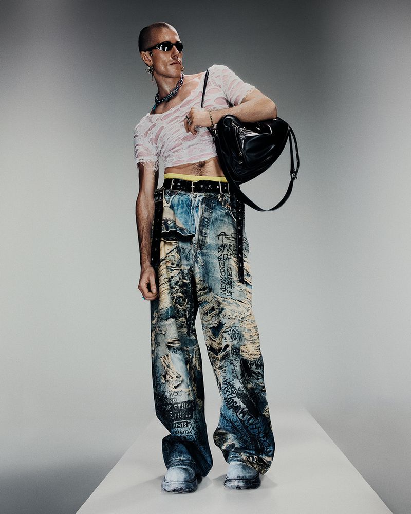 Fashionable adult male poses in ripped tee, printed jeans, white sneakers, and sunglasses against a gradient gray backdrop.