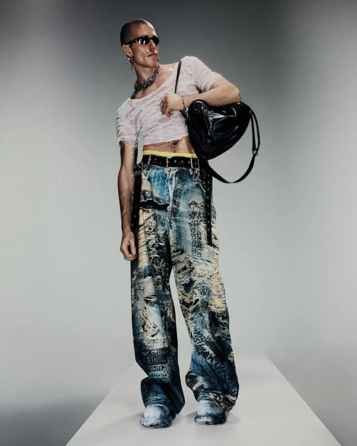 Fashionable adult male poses in ripped tee, printed jeans, white sneakers, and sunglasses against a gradient gray backdrop.