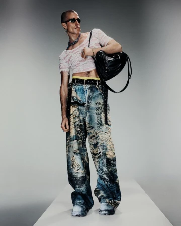 Fashionable adult male poses in ripped tee, printed jeans, white sneakers, and sunglasses against a gradient gray backdrop.