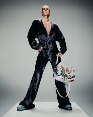 Model wearing a dark patterned jumpsuit holding a pale pink handbag against a grey studio seamless background