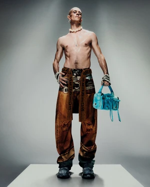 Shirtless male model wearing patterned brown trousers holding a bright blue handbag against a grey studio backdrop