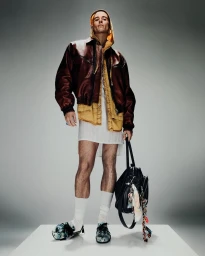 Model Jordan Robson in Acne Studios S/S 25 menswear, wearing a brown leather jacket and yellow hoodie over a white tunic, holding a black bag.
