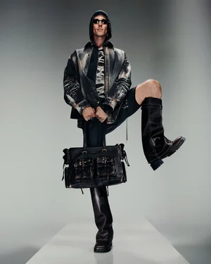 Model in hooded patterned jacket and tall boots holding a black leather bag for Acne Studios Spring/Summer 25 Men's Lookbook.
