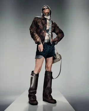 Model wearing patterned jacket, dark shorts, and tall leather boots against grey studio background