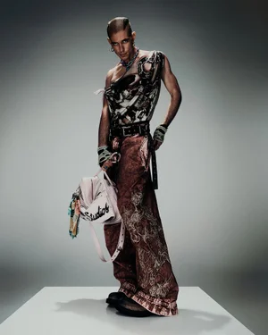 Model standing on a white pedestal wearing an asymmetrical printed top, patterned trousers, and holding a pink bag against a grey gradient background