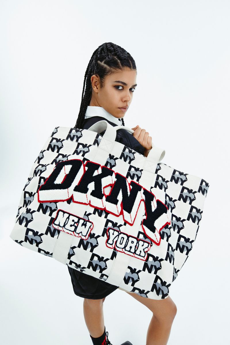 Fashion shot of a young woman carrying a DKNY New York logo tote bag against a bright white backdrop in a studio setting.