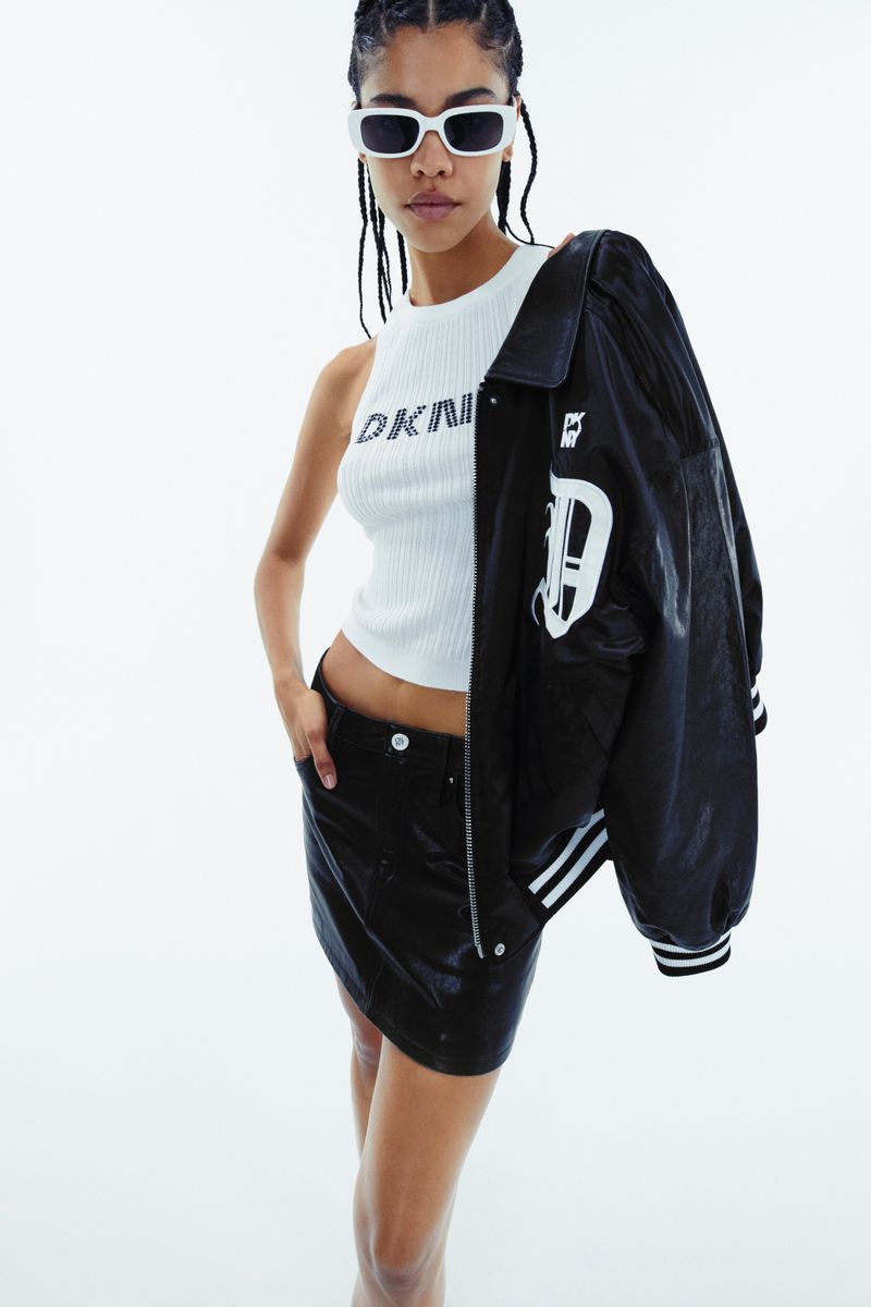 A fashionable young woman in a DKNY top and black miniskirt poses against a white background, wearing sunglasses and a varsity jacket.