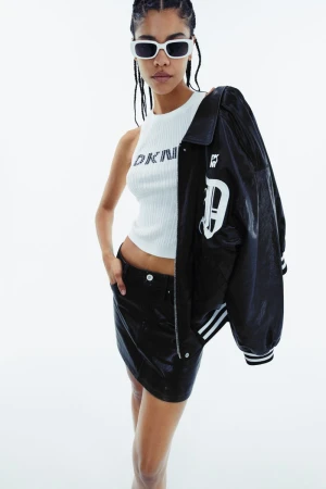 A fashionable young woman in a DKNY top and black miniskirt poses against a white background, wearing sunglasses and a varsity jacket.