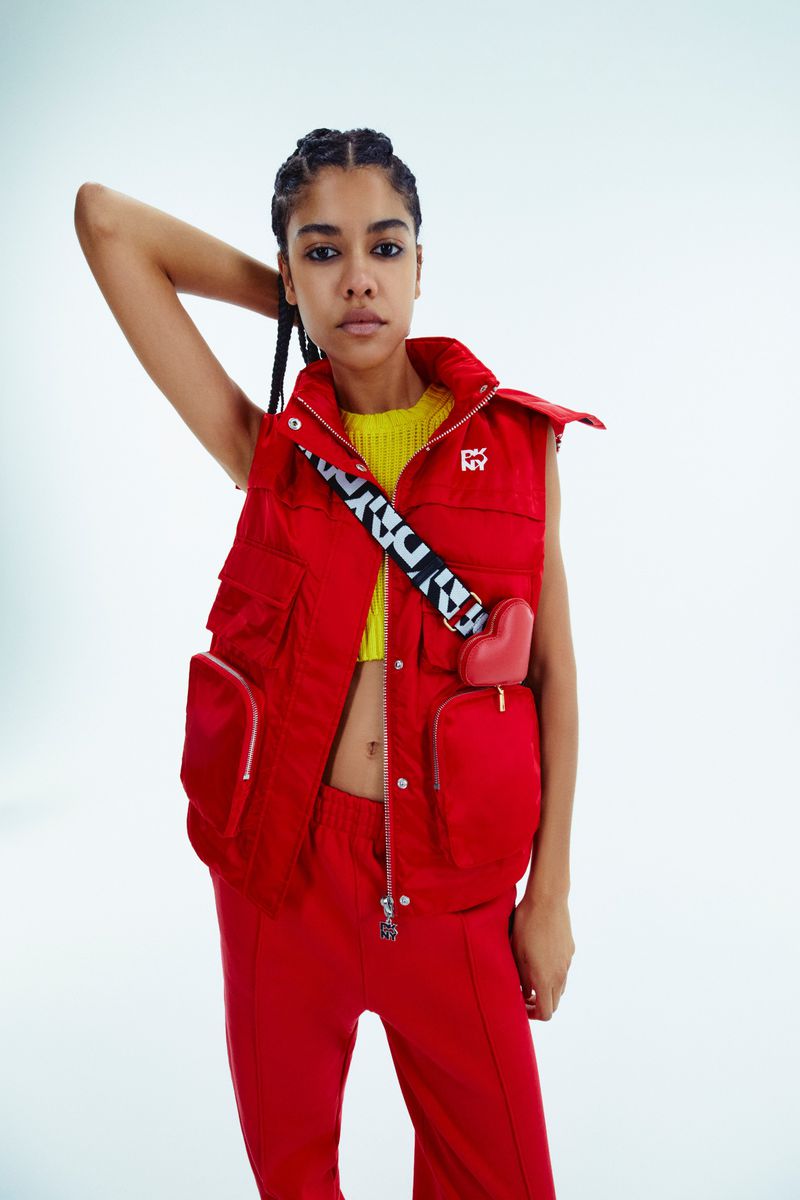 A woman poses wearing a vibrant yellow top, red vest, red pants, and a black-and-white strap, set against a clean white backdrop.