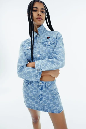 Young woman in a patterned denim jacket and mini skirt poses in a photo studio. The look is completed with long braids.
