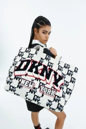 Model carrying a large printed canvas tote bag with bold DKNY logo against a plain white studio background.