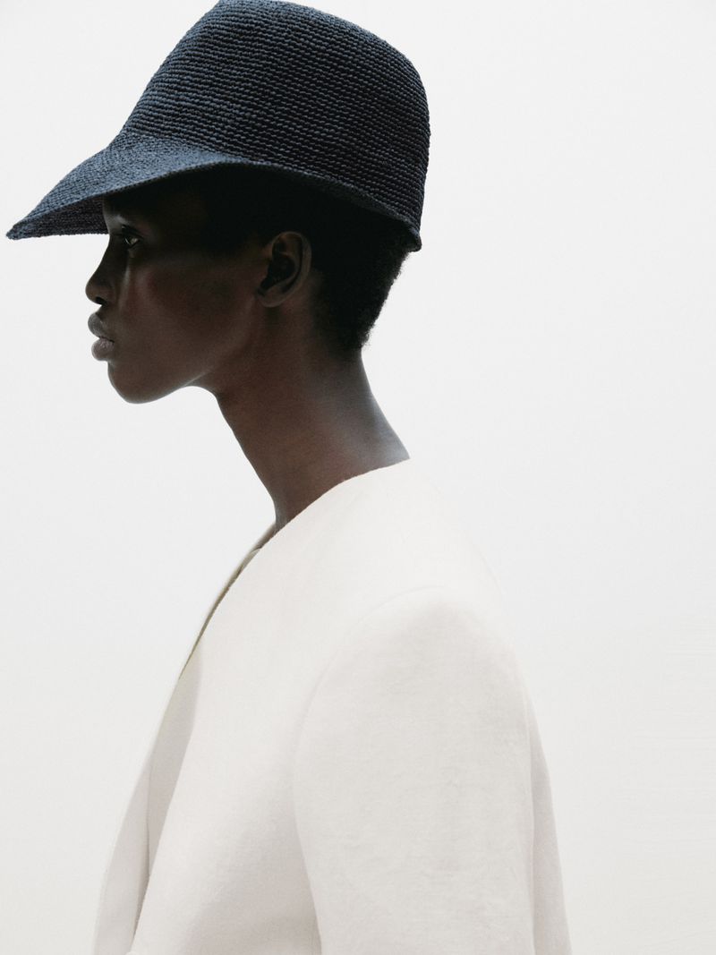 Profile shot of a woman with dark skin wearing a dark woven hat and a light-colored blazer against a clean white background.