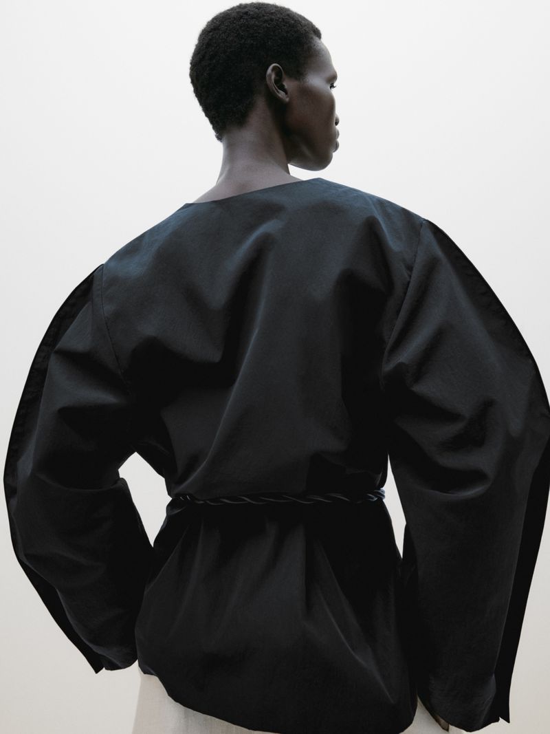 Back view of an African-descent adult female posing in a draped, black top cinched at the waist with a braided belt on a white background.