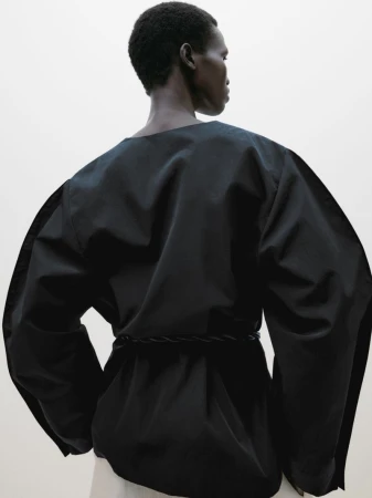 Back view of an African-descent adult female posing in a draped, black top cinched at the waist with a braided belt on a white background.