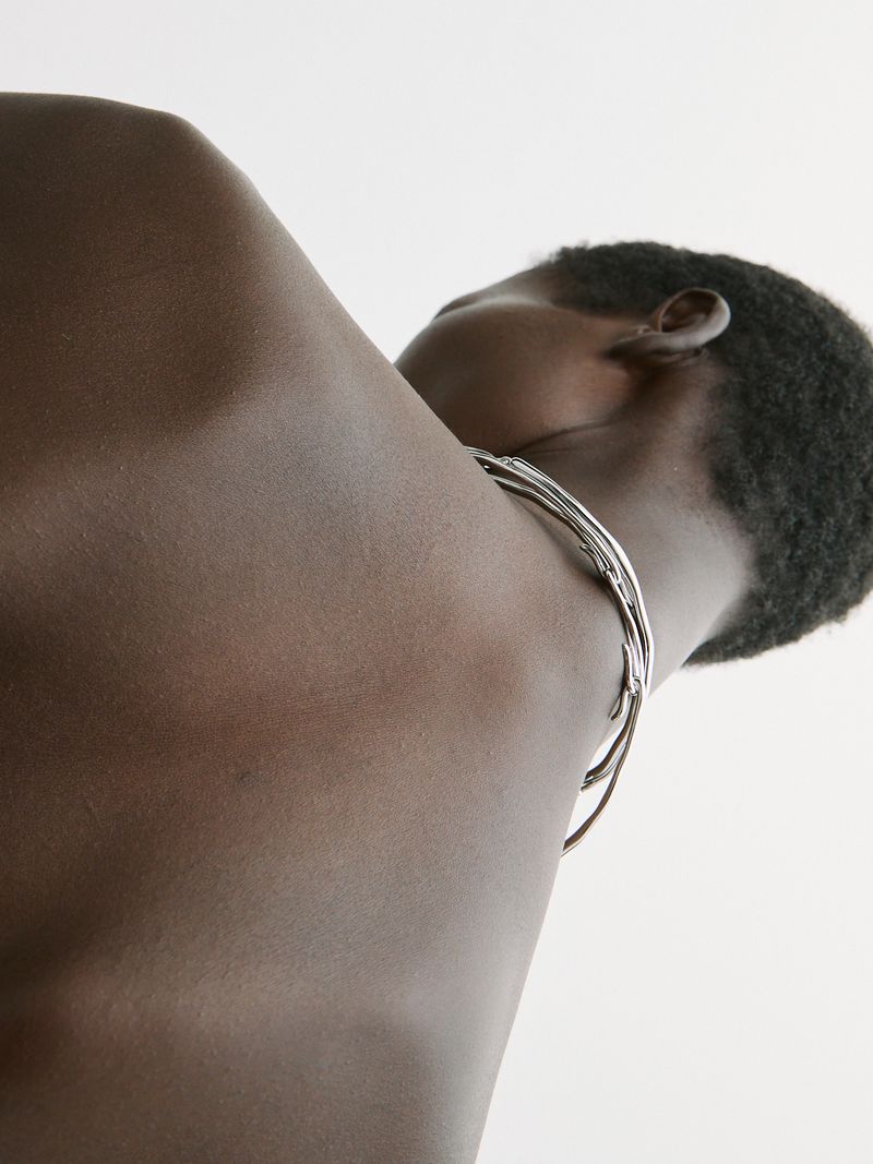 Close-up on an adult with dark skin and short hair, showcasing a silver metal neck choker against a plain white background.