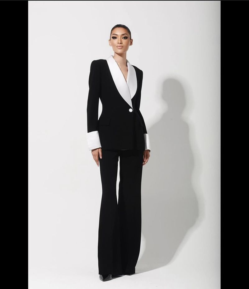 Elegant woman in a black suit with a white collar and cuffs posing in a studio with a bright background, showcasing fashion attire.