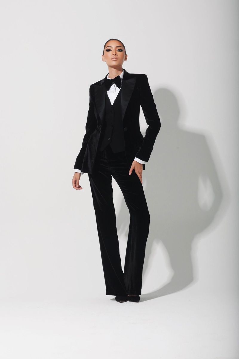 A woman in a black velvet tuxedo suit, vest, and bow tie strikes a pose against a white backdrop in a fashion portrait.