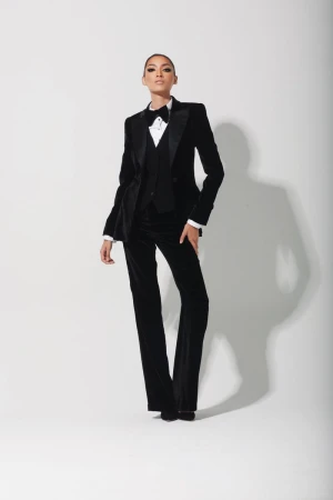 A woman in a black velvet tuxedo suit, vest, and bow tie strikes a pose against a white backdrop in a fashion portrait.