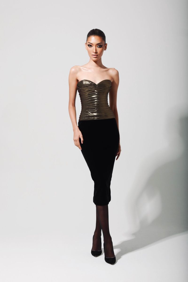 A woman stands in a photo studio wearing a gold bustier top, a black pencil skirt, stockings, and pumps against a solid white backdrop.