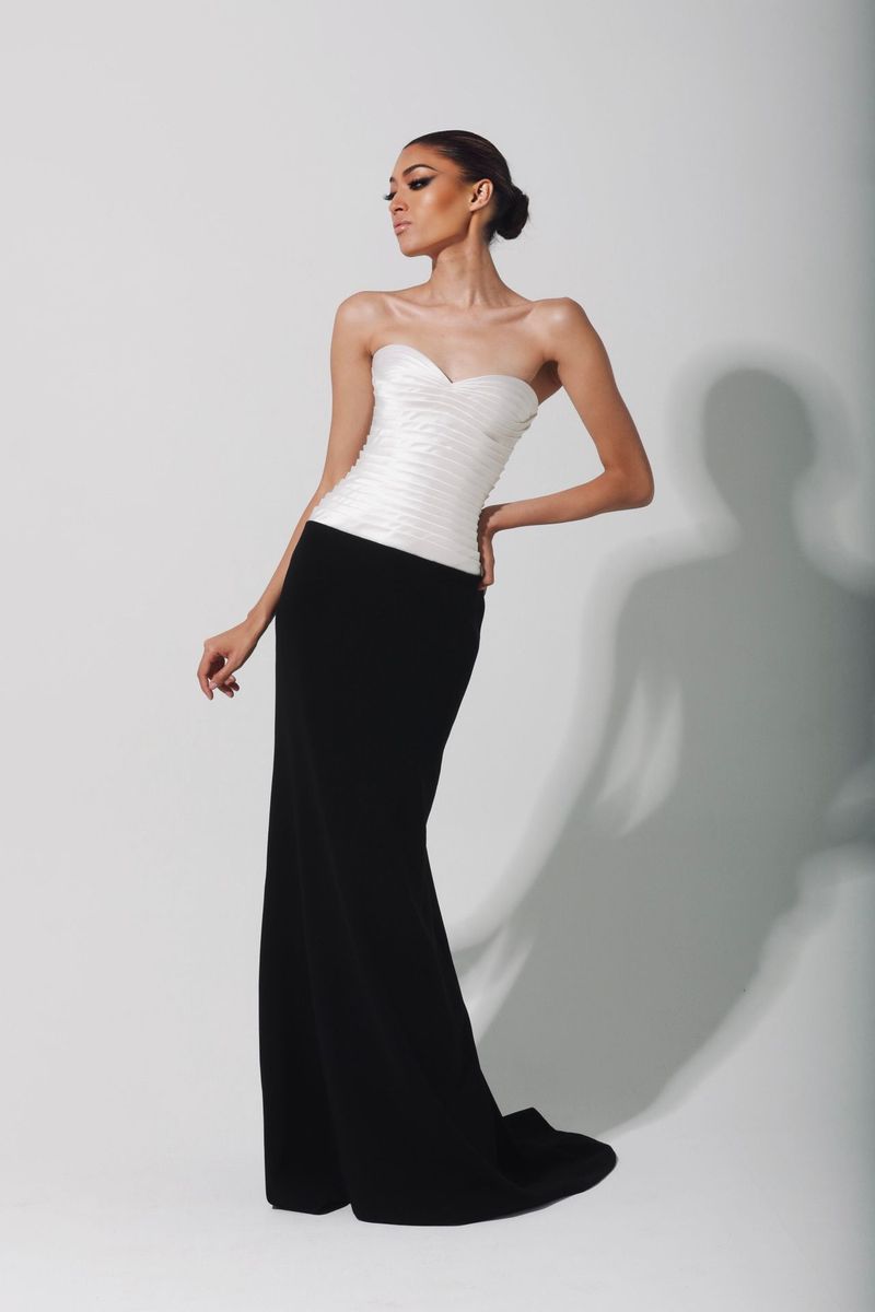 A woman poses in a floor-length, strapless evening gown with a white ruched bodice and black skirt against a neutral background.