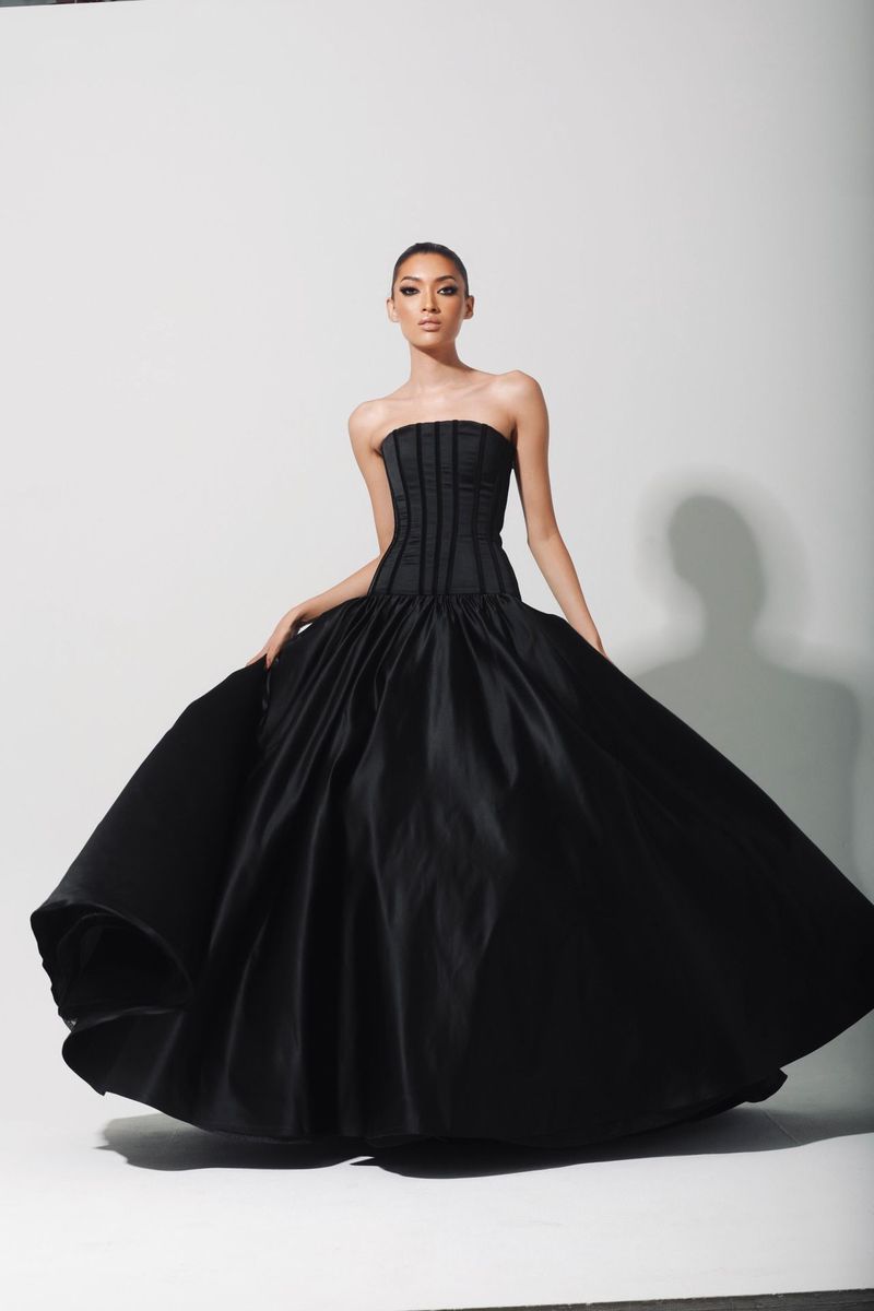 A woman poses in a black strapless gown with a full skirt against a white backdrop, showcasing a fashion-forward and elegant style.
