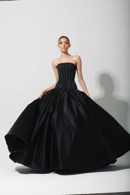A woman poses in a black strapless gown with a full skirt against a white backdrop, showcasing a fashion-forward and elegant style.