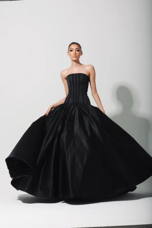 A woman poses in a black strapless gown with a full skirt against a white backdrop, showcasing a fashion-forward and elegant style.