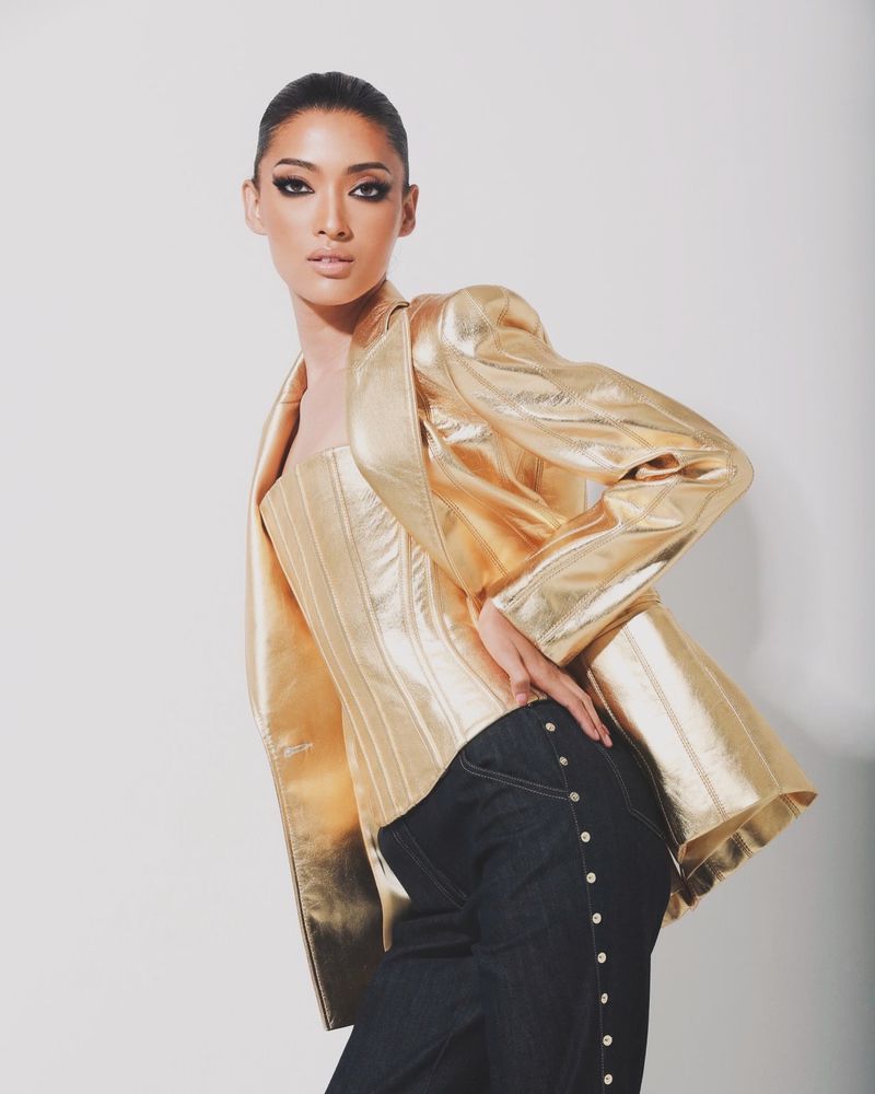Woman in gold corset and jacket poses against a white backdrop, showcasing a stylish and modern fashion aesthetic.