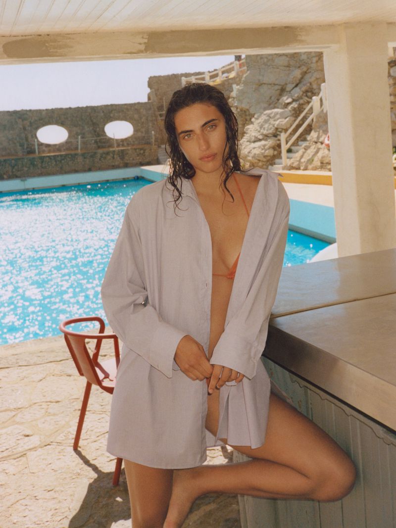 Adult female with wet hair poses in an oversized shirt near a turquoise pool, stone structure, and red chair in an outdoor setting.